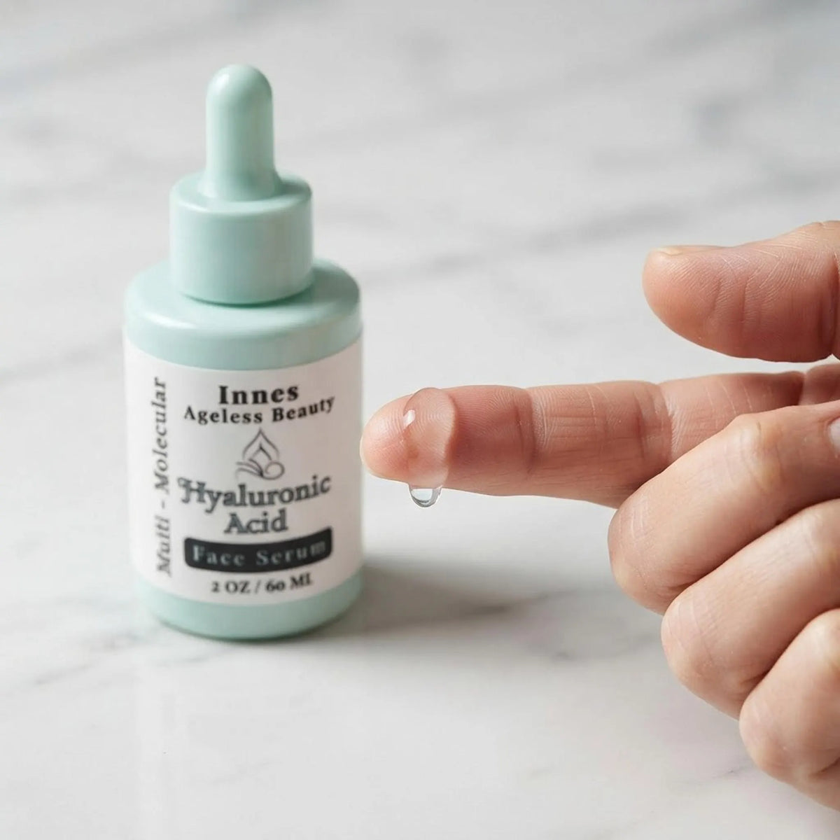 Macro shot of a clear drop of facial serum on a fingertip next to the Innes Ageless Beauty serum bottle.