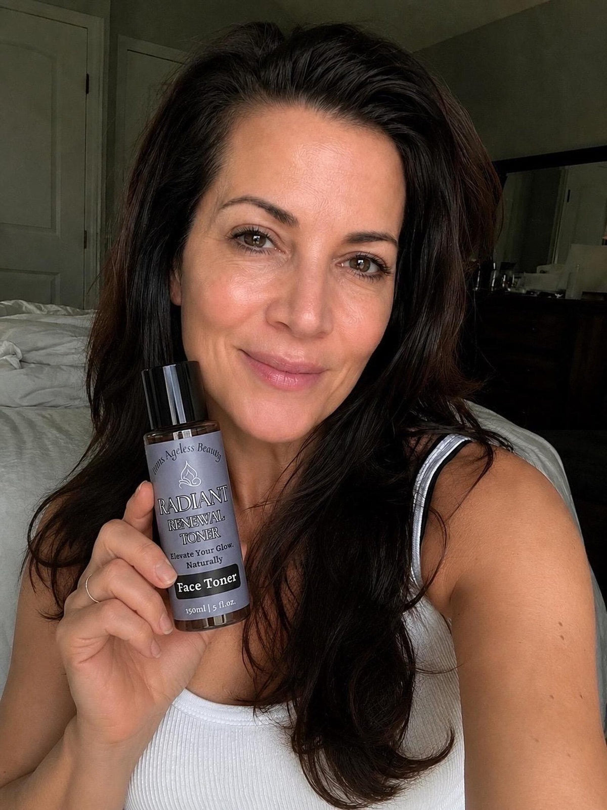 A smiling woman with long dark hair holds a clear spray bottle labeled Radiant Renewal Toner Face Toner.