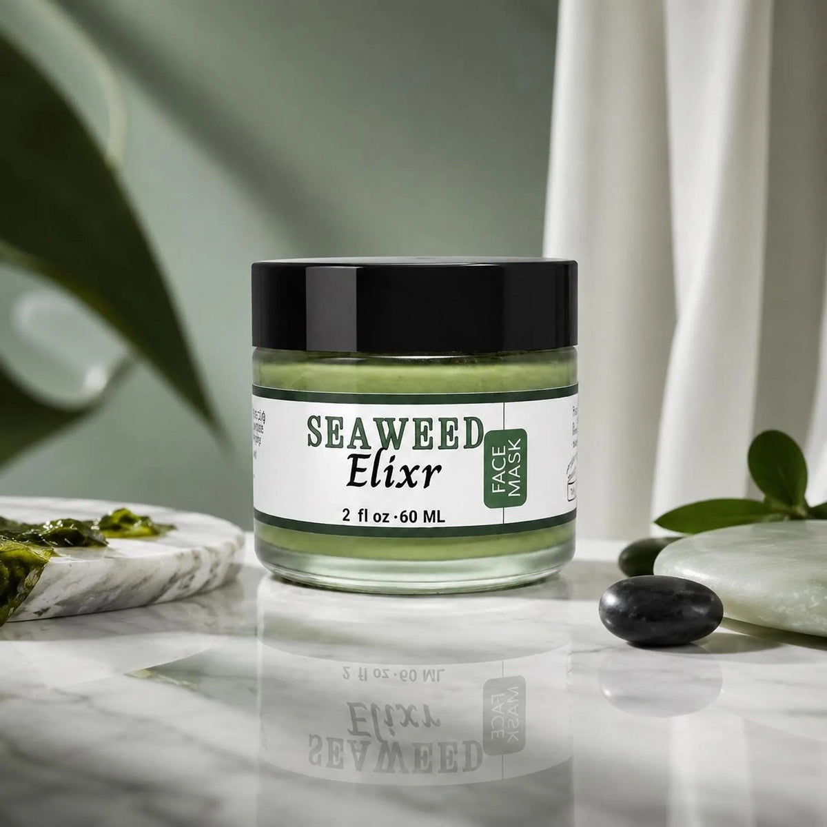 A glass jar of green seaweed face mask with a black lid sits on a reflective white marble surface next to smooth pebbles and green leaves.