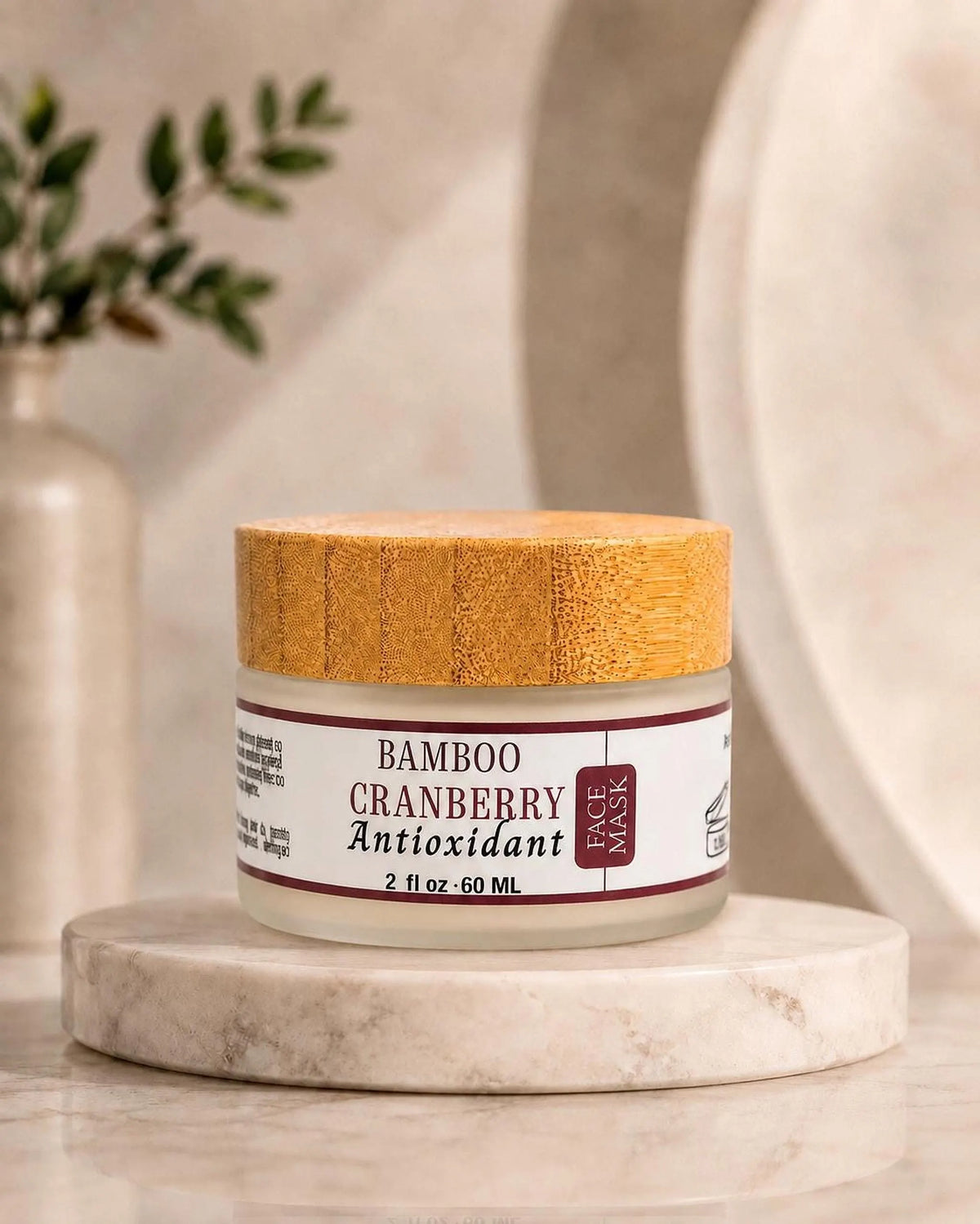 The jar of Bamboo Cranberry Antioxidant face mask stands on a round marble platform against a blurred neutral background with a vase of greenery.