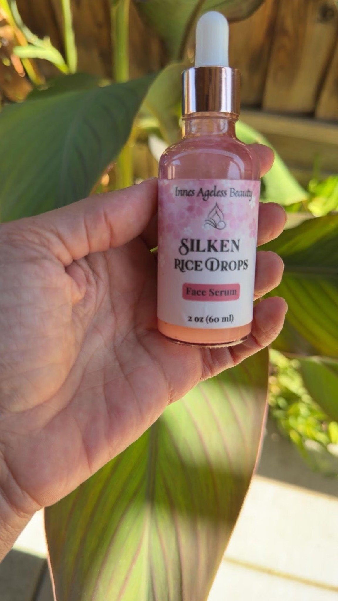 Hand holding a 2 oz bottle of Silken Rice Drops face serum against sunlit tropical green leaves.