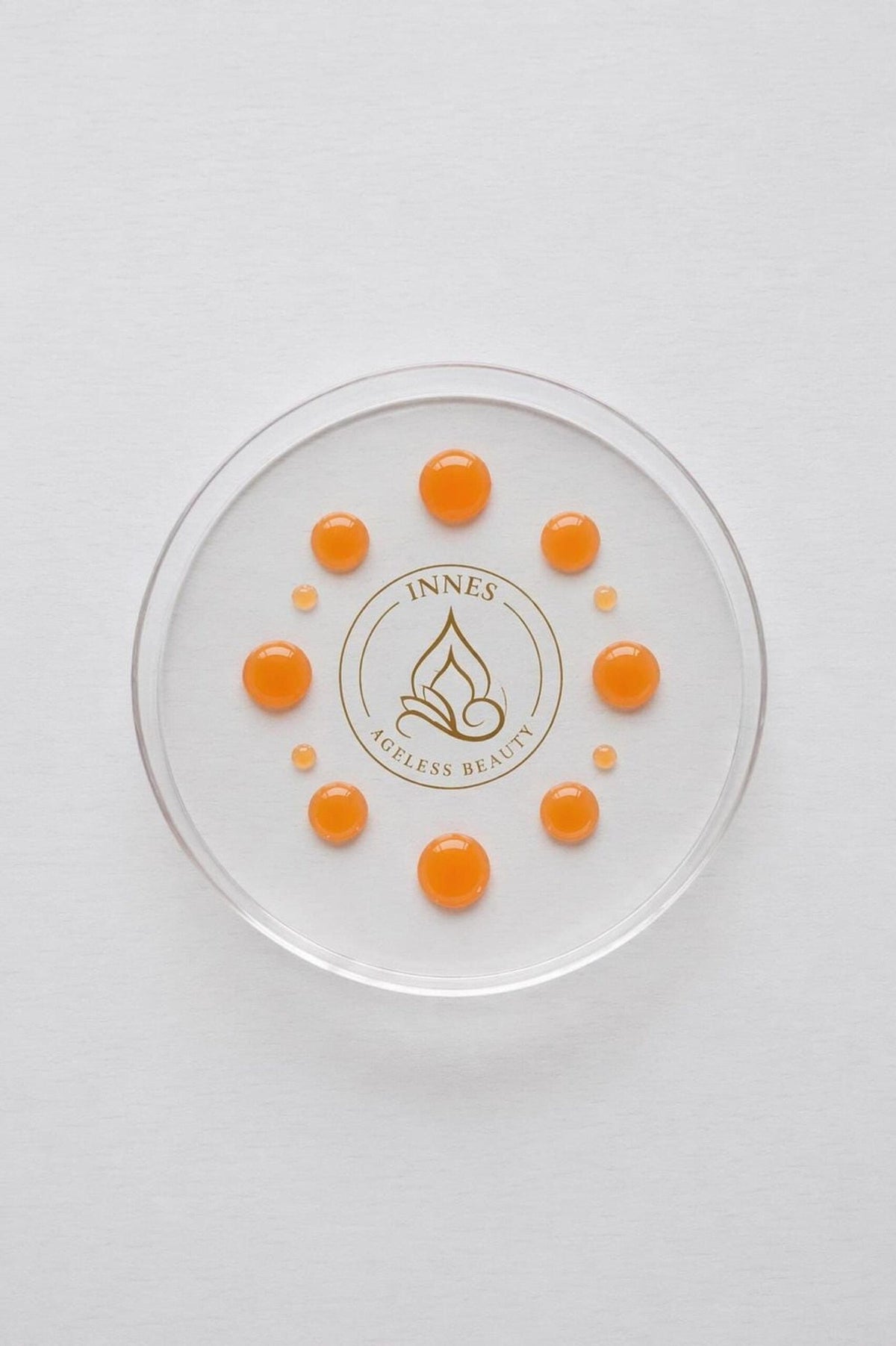 A clear circular glass dish containing multiple orange serum droplets arranged in a circle over a white textured background, with the Innes Ageless Beauty logo printed in the center.