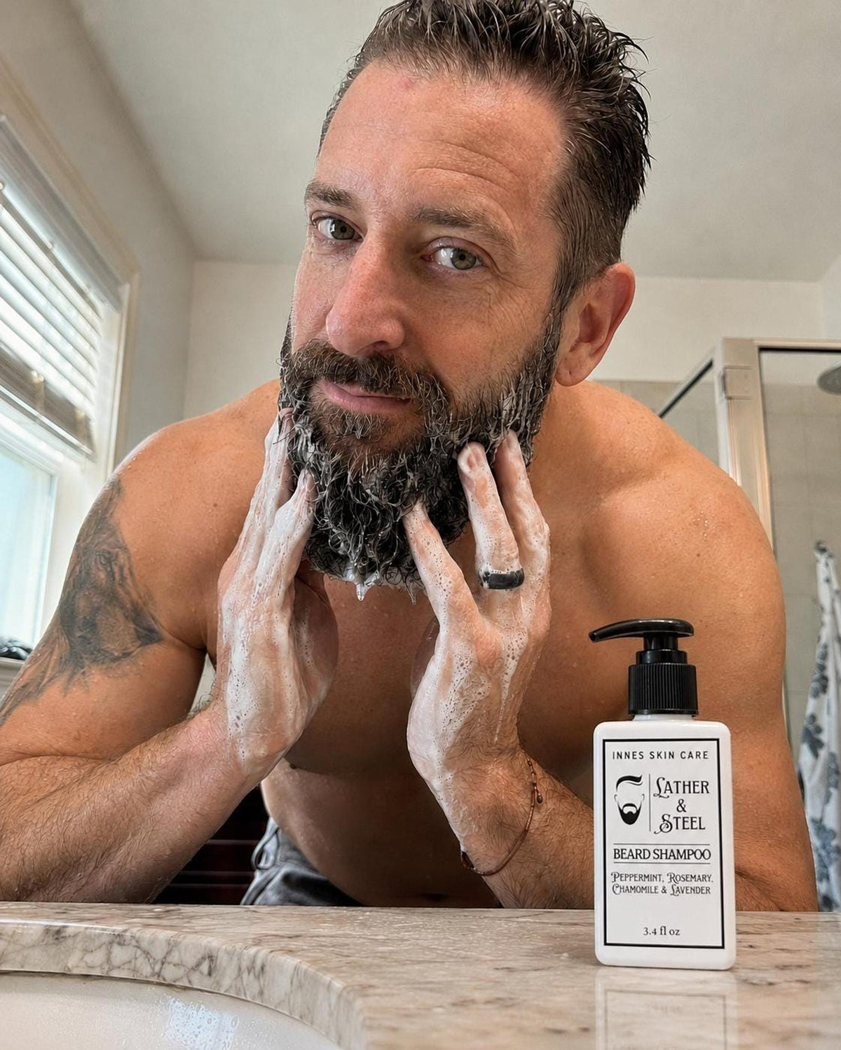 A shirtless man with a tattoo on his arm lathers white foam into his beard next to the bottle of beard shampoo on a counter.