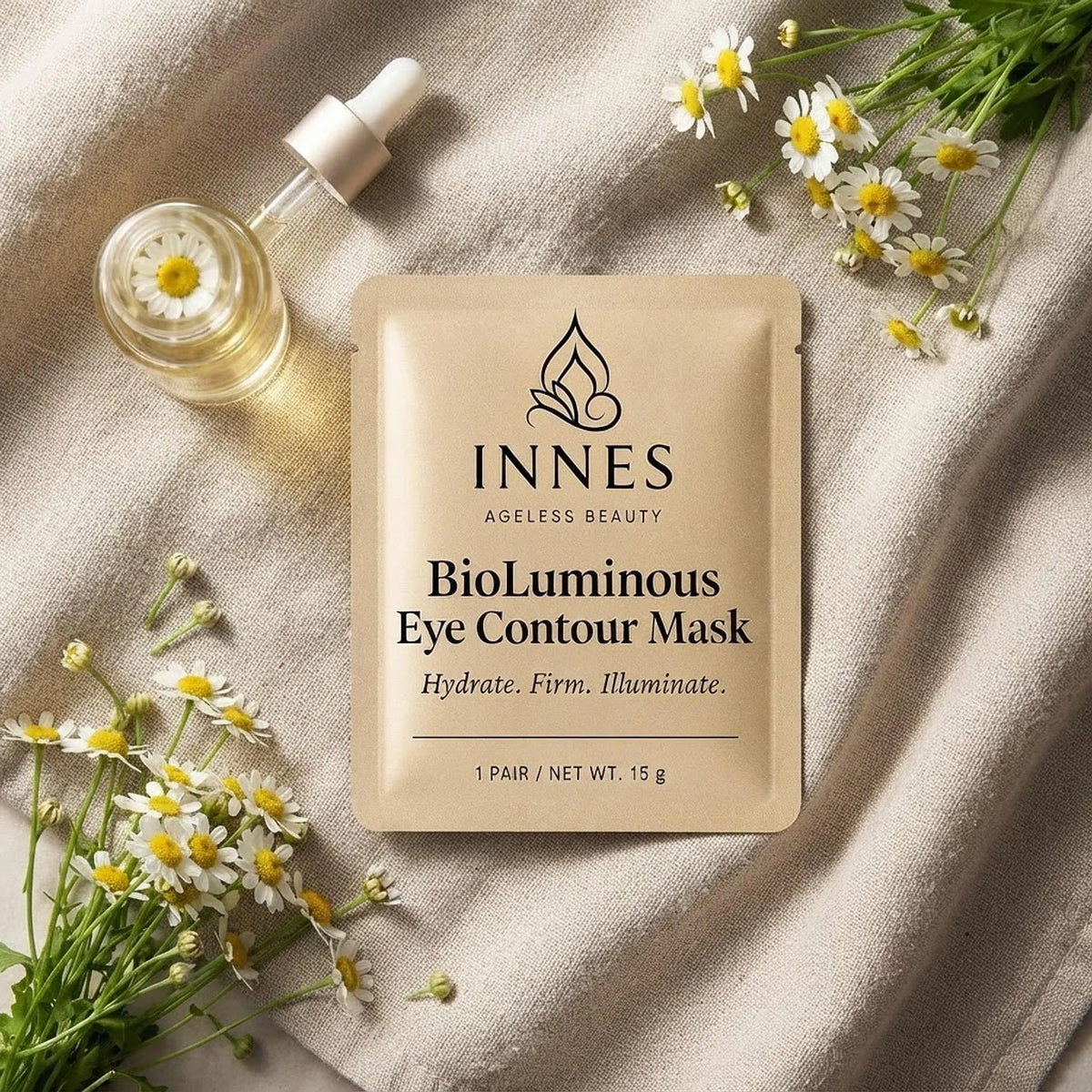 Overhead flat lay of the gold Innes BioLuminous Eye Contour Mask pouch on beige linen with chamomile flowers and serum.