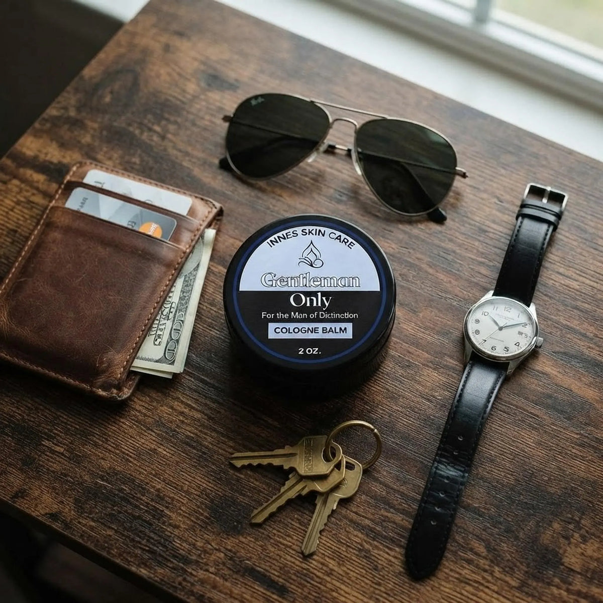 The 2 oz black cologne balm jar laid out flat with a wallet, watch, and sunglasses on a wooden table.