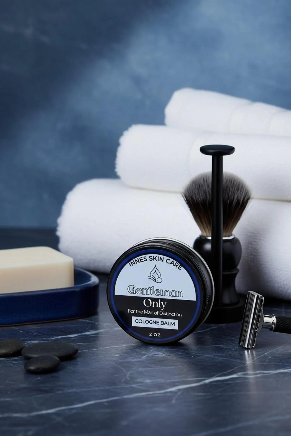 A black cologne balm tin with the text "Gentleman Only" sits on a dark surface. A shaving brush, razor, soap, and black stones are also present. White towels are stacked in the background.