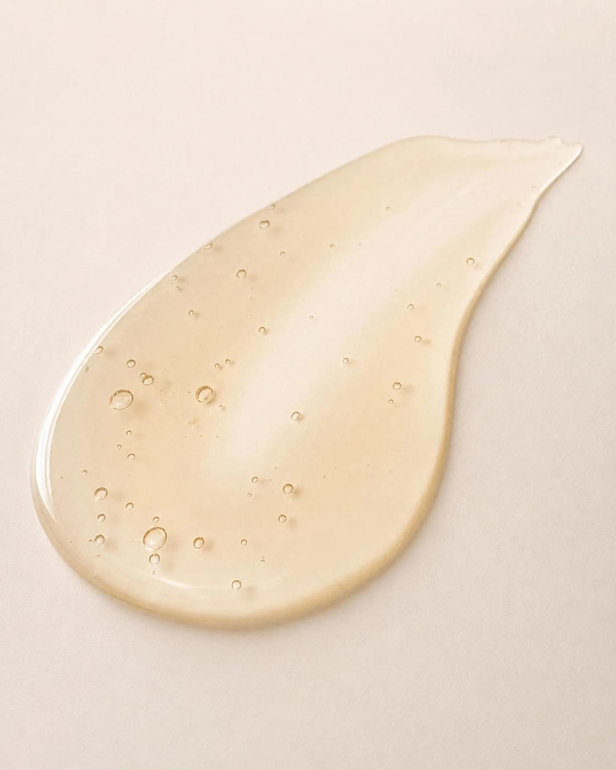 A glossy, translucent yellow-gold droplet of serum with tiny air bubbles scattered throughout, smeared on a neutral light beige surface.