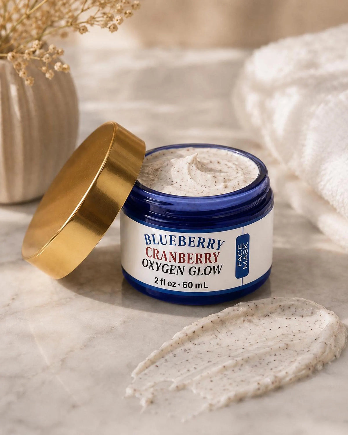 A blue jar of Blueberry Cranberry Oxygen Glow face mask with its gold lid open, showing creamy speckled product next to a smear on a marble surface.