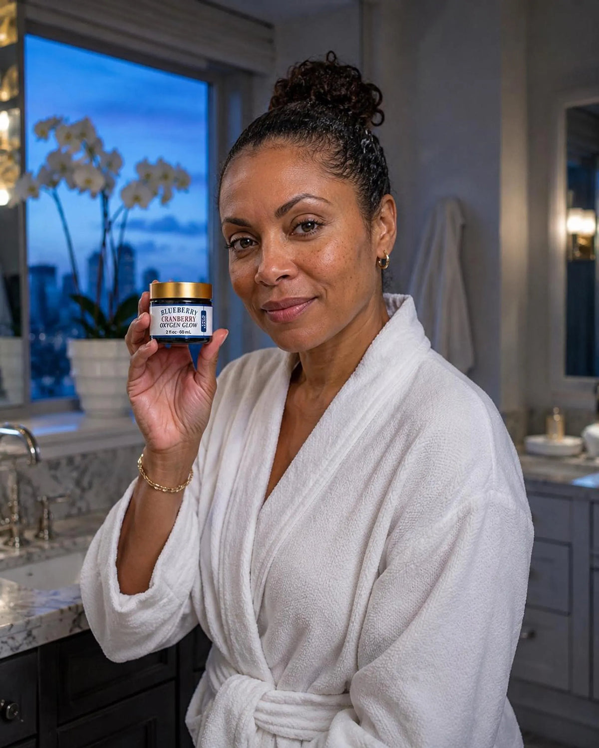 A woman wearing a white bathrobe holds up the blue jar of face mask in a bathroom setting at twilight.