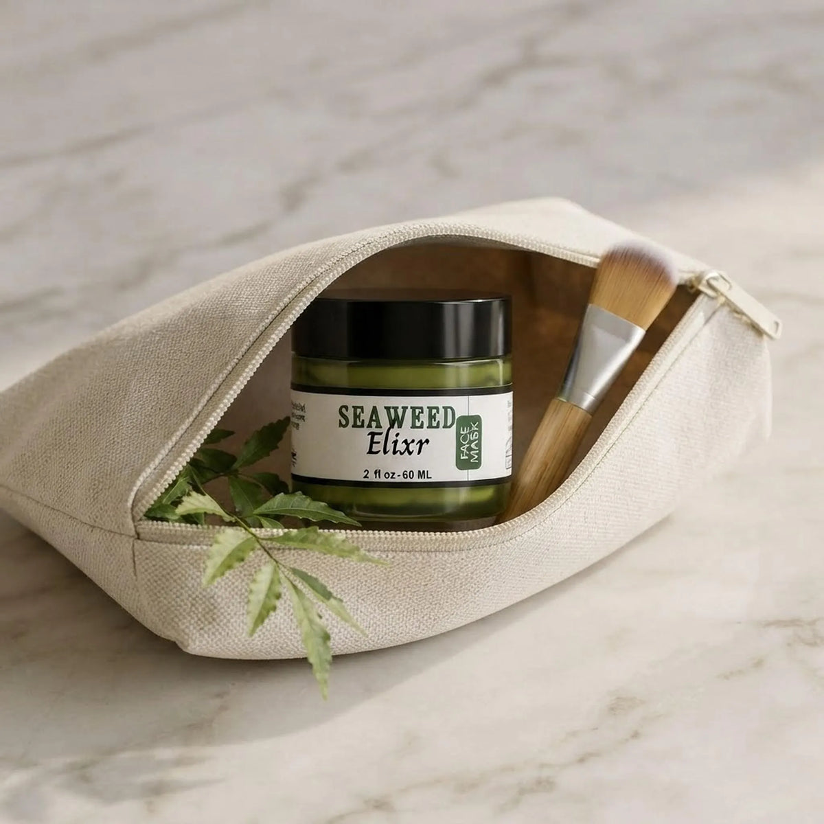 A glass jar of green seaweed face mask with a black lid sits on a reflective white marble surface next to smooth pebbles and green leaves.