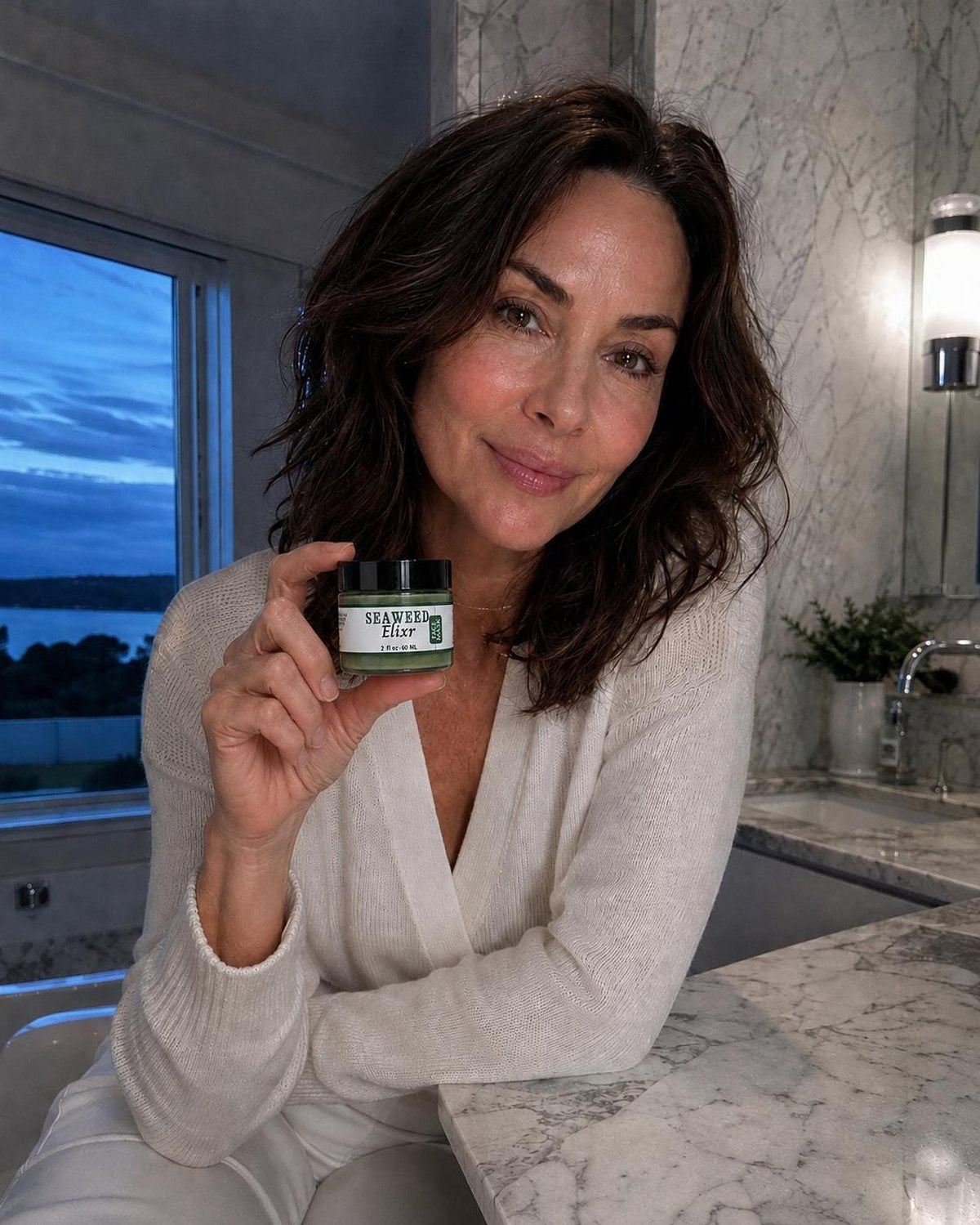 A woman with dark hair wearing a white cardigan holds a small green jar labeled Seaweed Elixir while sitting in a bathroom with marble countertops.