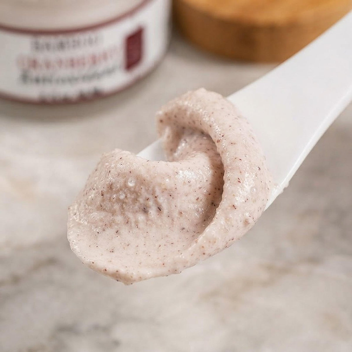 An open jar of Bamboo Cranberry Antioxidant face mask with a wooden lid sits on a marble surface next to a swatch of the pink exfoliating cream.