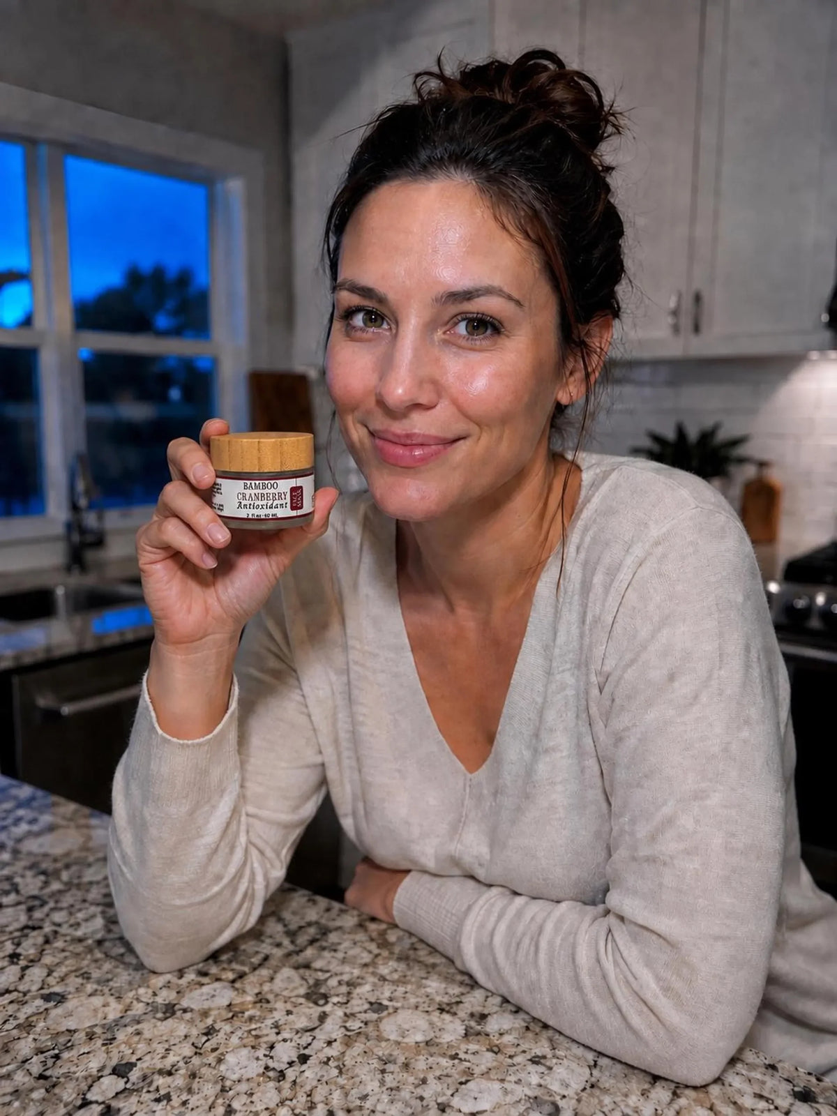 A woman smiles while holding the jar of Bamboo Cranberry Antioxidant face mask in a kitchen setting.